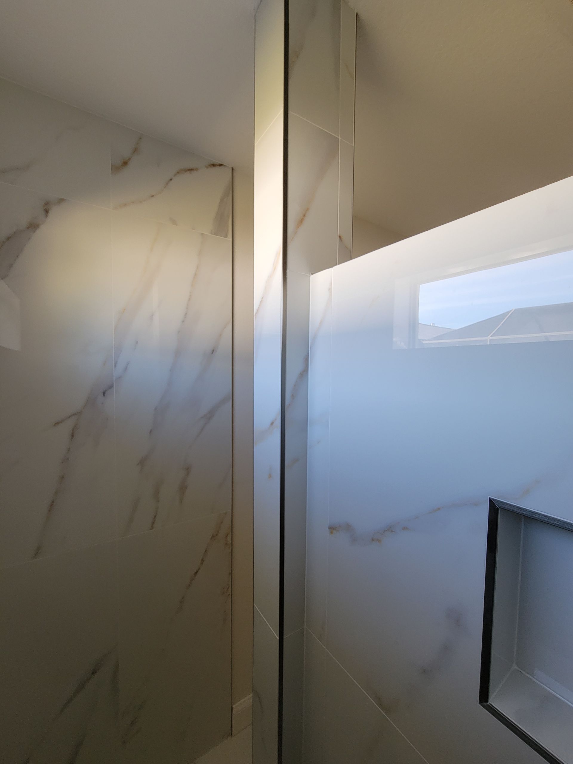Glass shower with marble-look tile walls. Narrow metal frame separates glass panels. Recessed shelf visible.