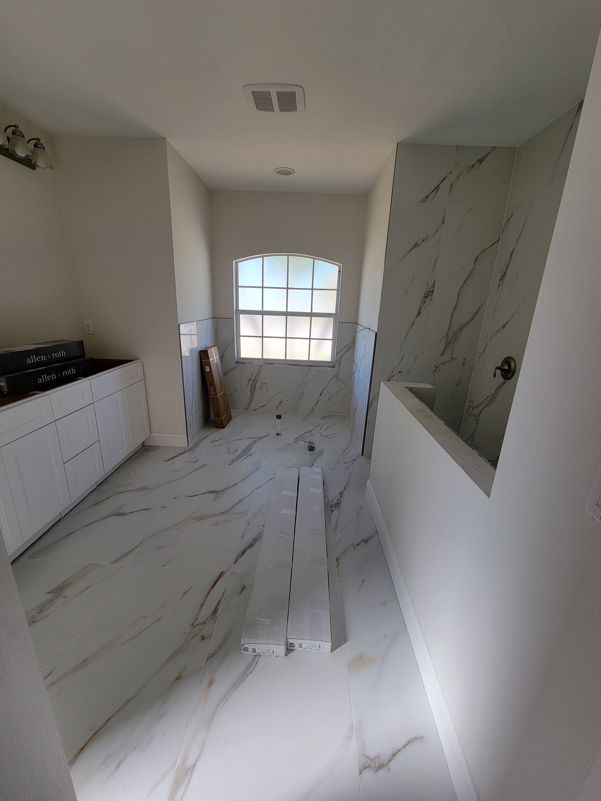 Bathroom under construction with marble-patterned tile and a window; white walls, unfinished cabinets.