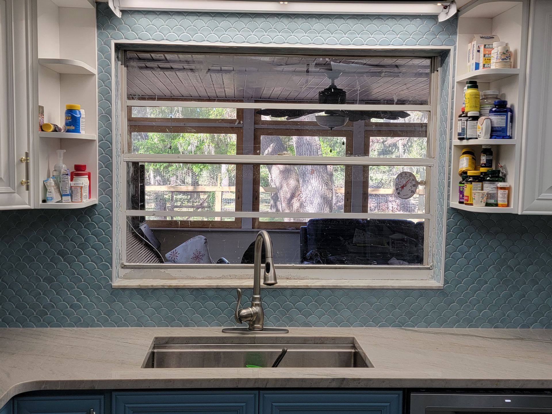 Kitchen with a window above a sink, blue cabinets, and white shelves. Green trees seen through the window.