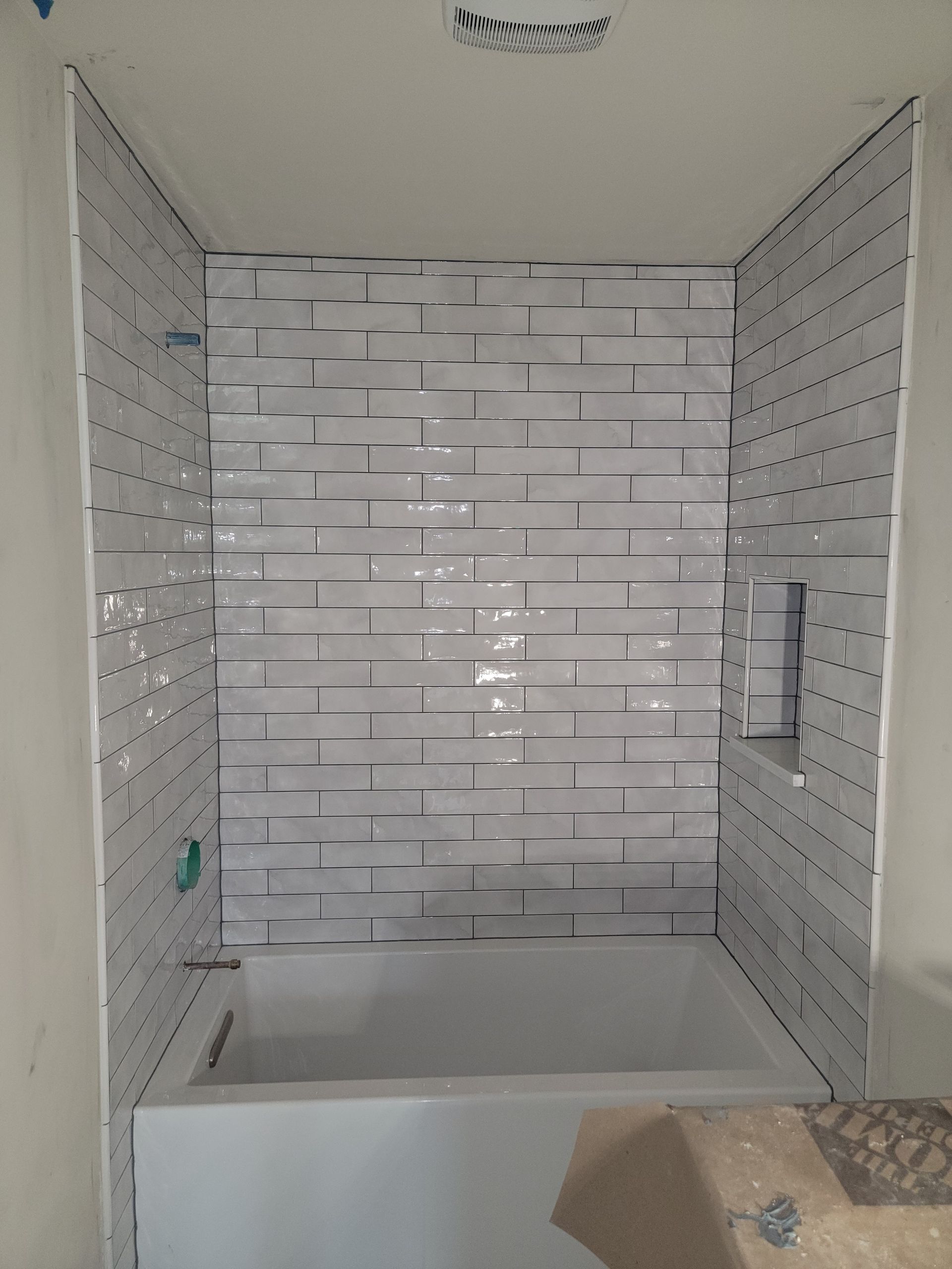 Tiled shower stall with white rectangular tiles and a built-in shelf. Tub at the bottom.