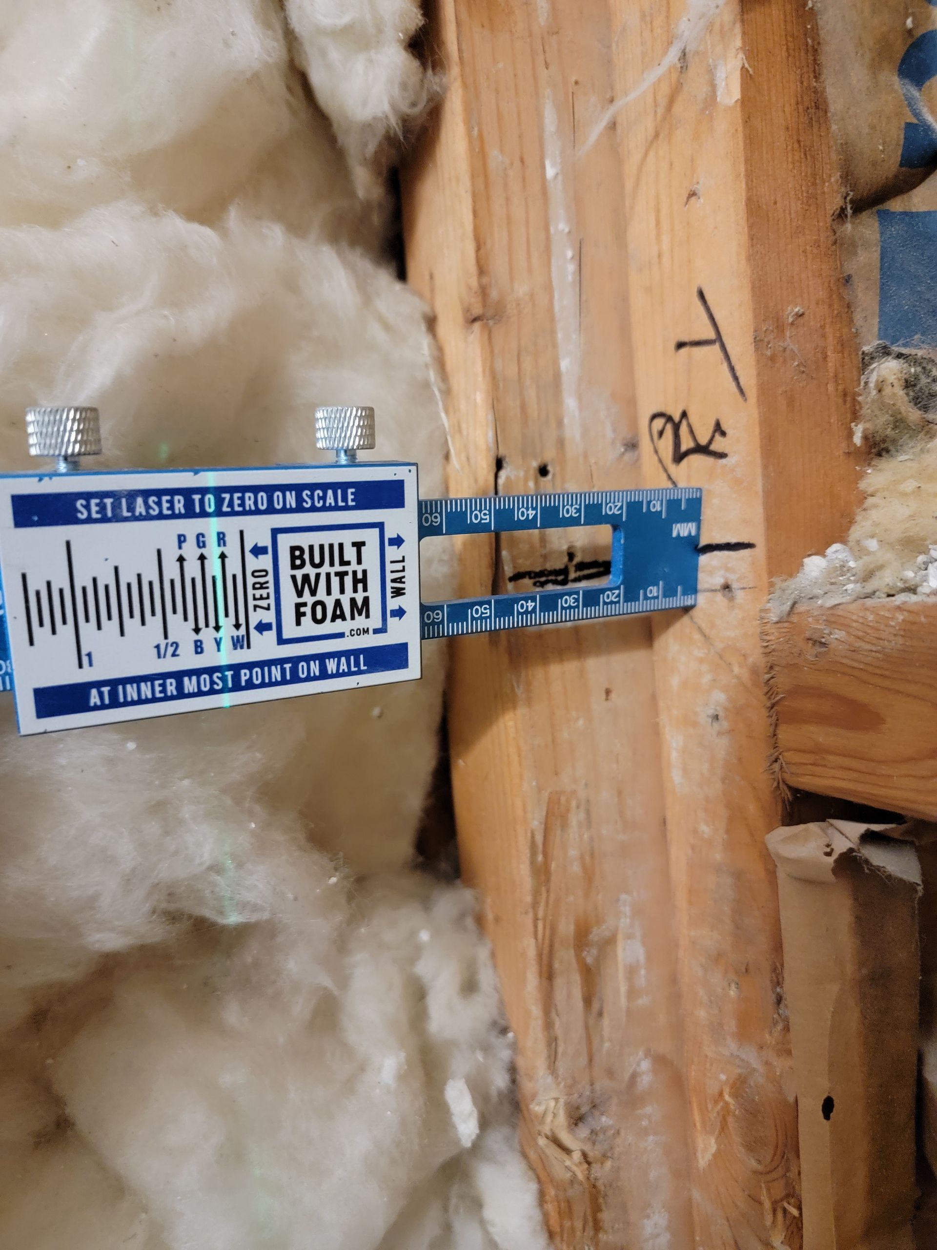 A blue measuring tool is against wood, next to insulation. The tool is labeled 