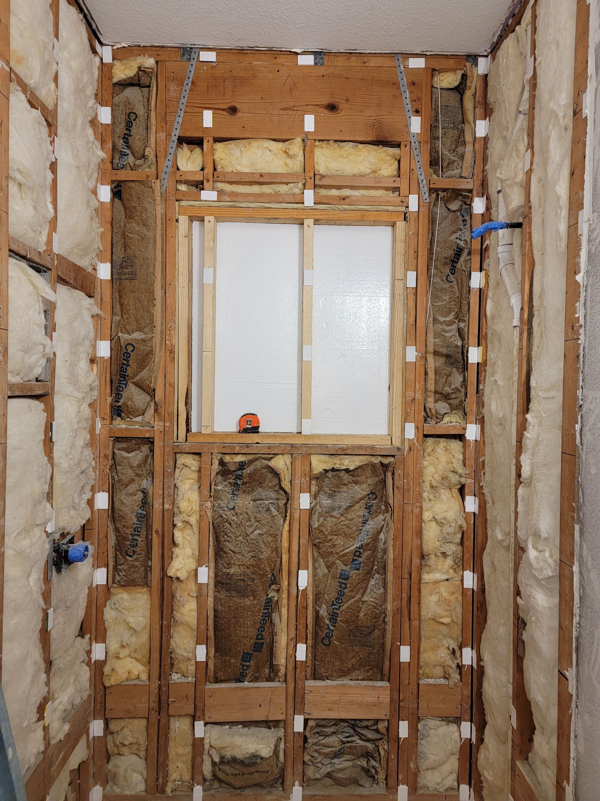 Bathroom renovation in progress, framing and insulation visible. Window centered, studs exposed, plumbing fixtures present.