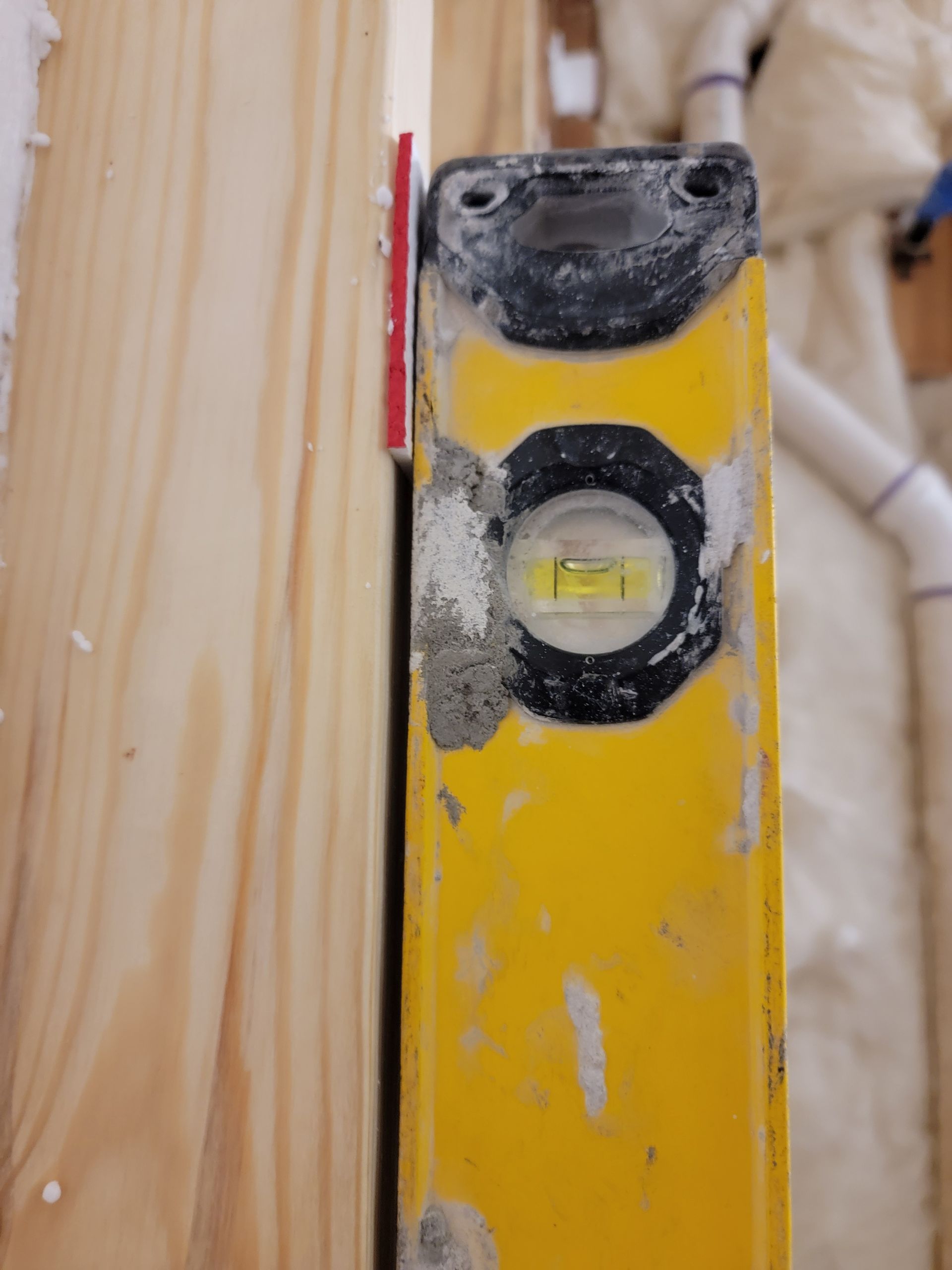 Yellow level pressed against a wooden stud with a red shim. Bubble in level is centered.