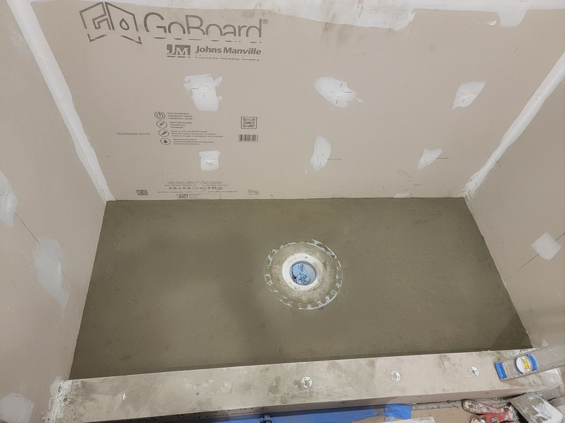 Shower construction in progress, gray cement floor with drain, light brown walls, GoBoard ceiling.