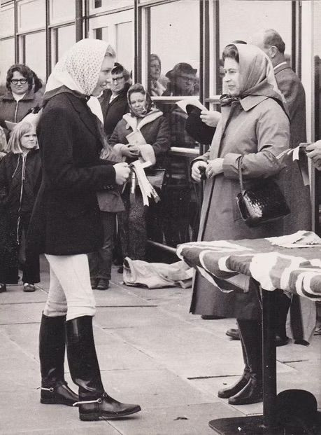 Two women stand facing each other outdoors, one holding papers, with a table and onlookers behind them.