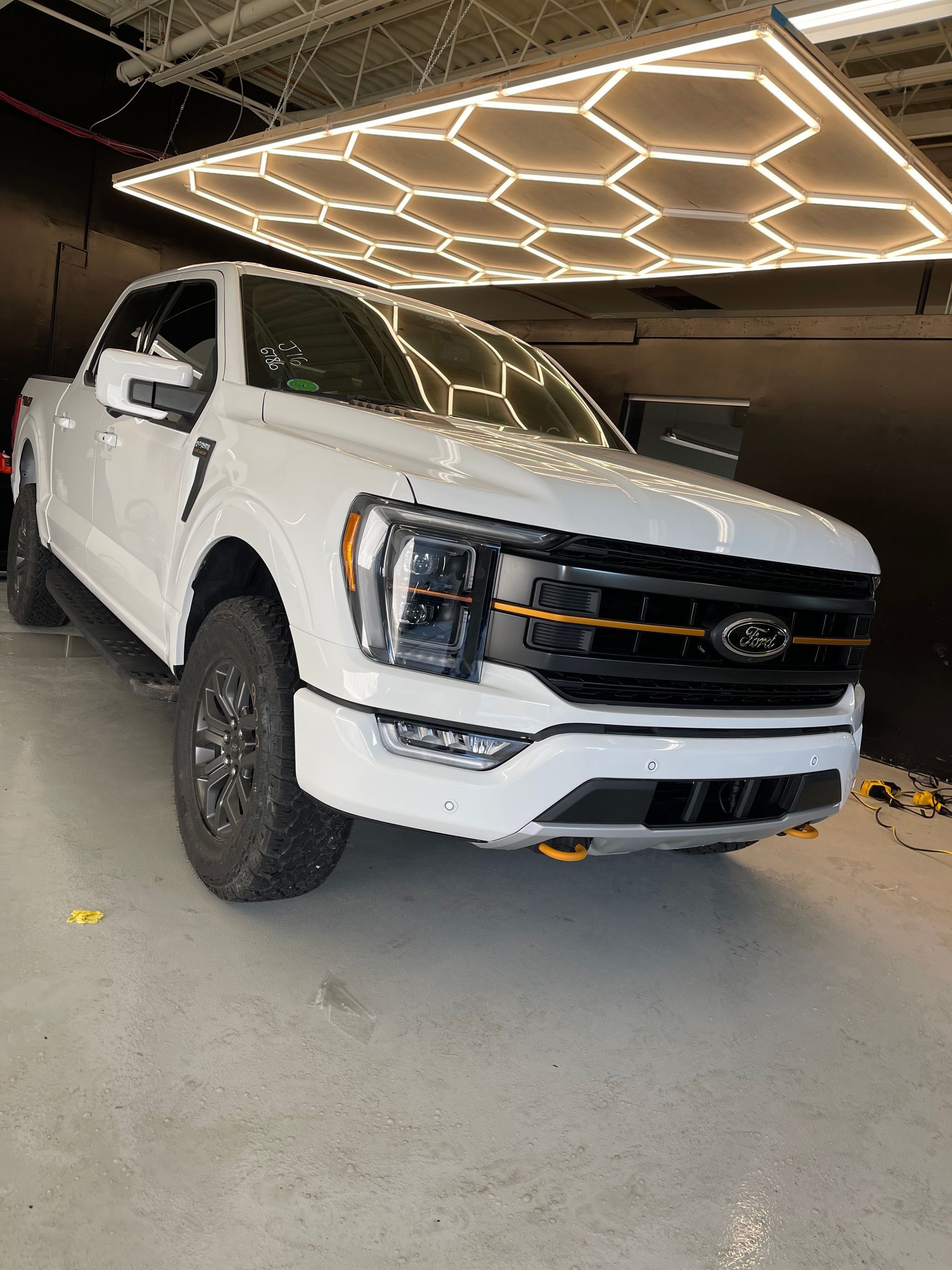 A white Ford F-150 pickup truck with black grille accents and orange tow hooks, parked under honeycomb ceiling lights.