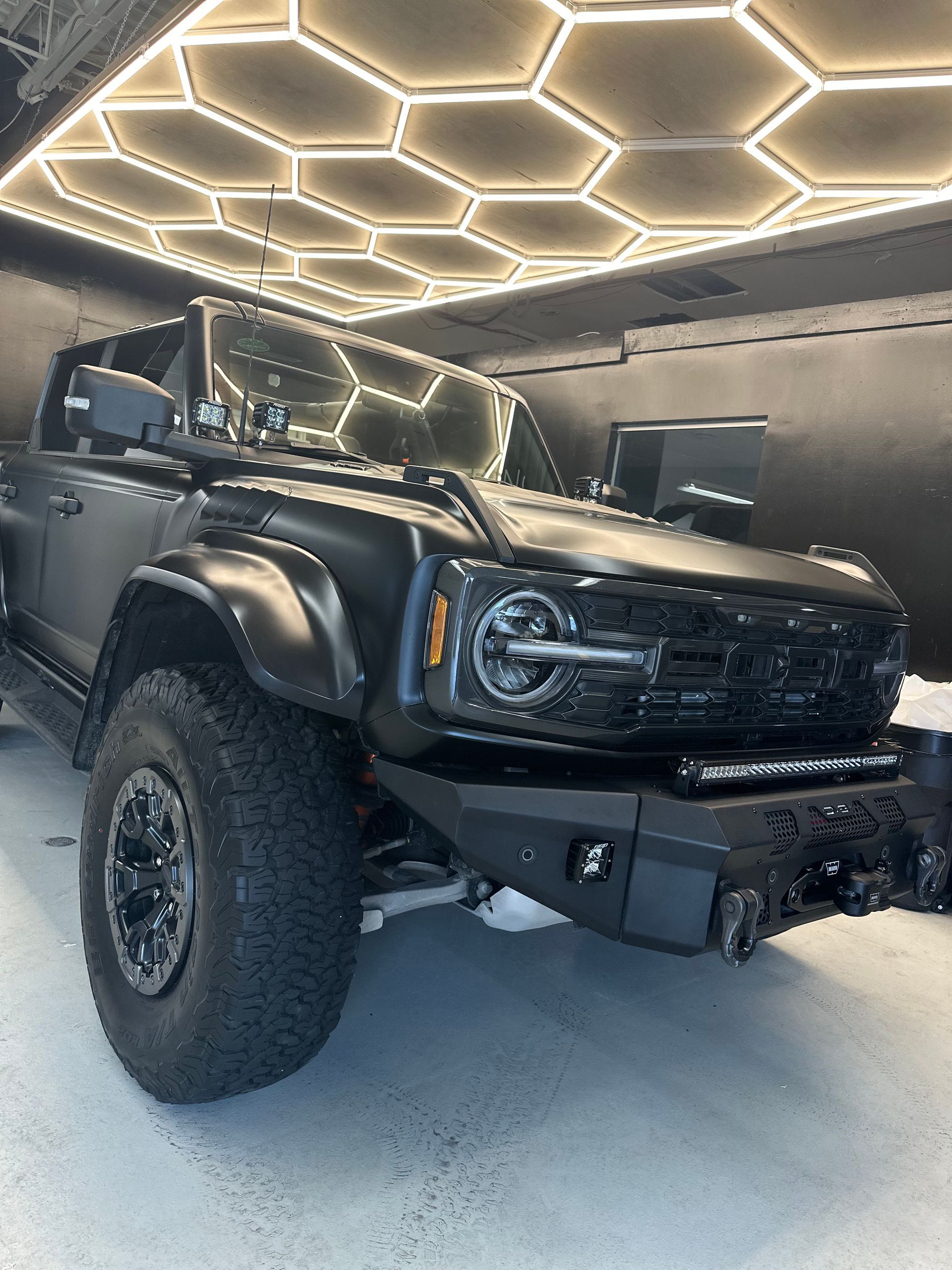 A matte black Ford Bronco with off-road modifications parked in a garage with a hexagonal LED ceiling light pattern.