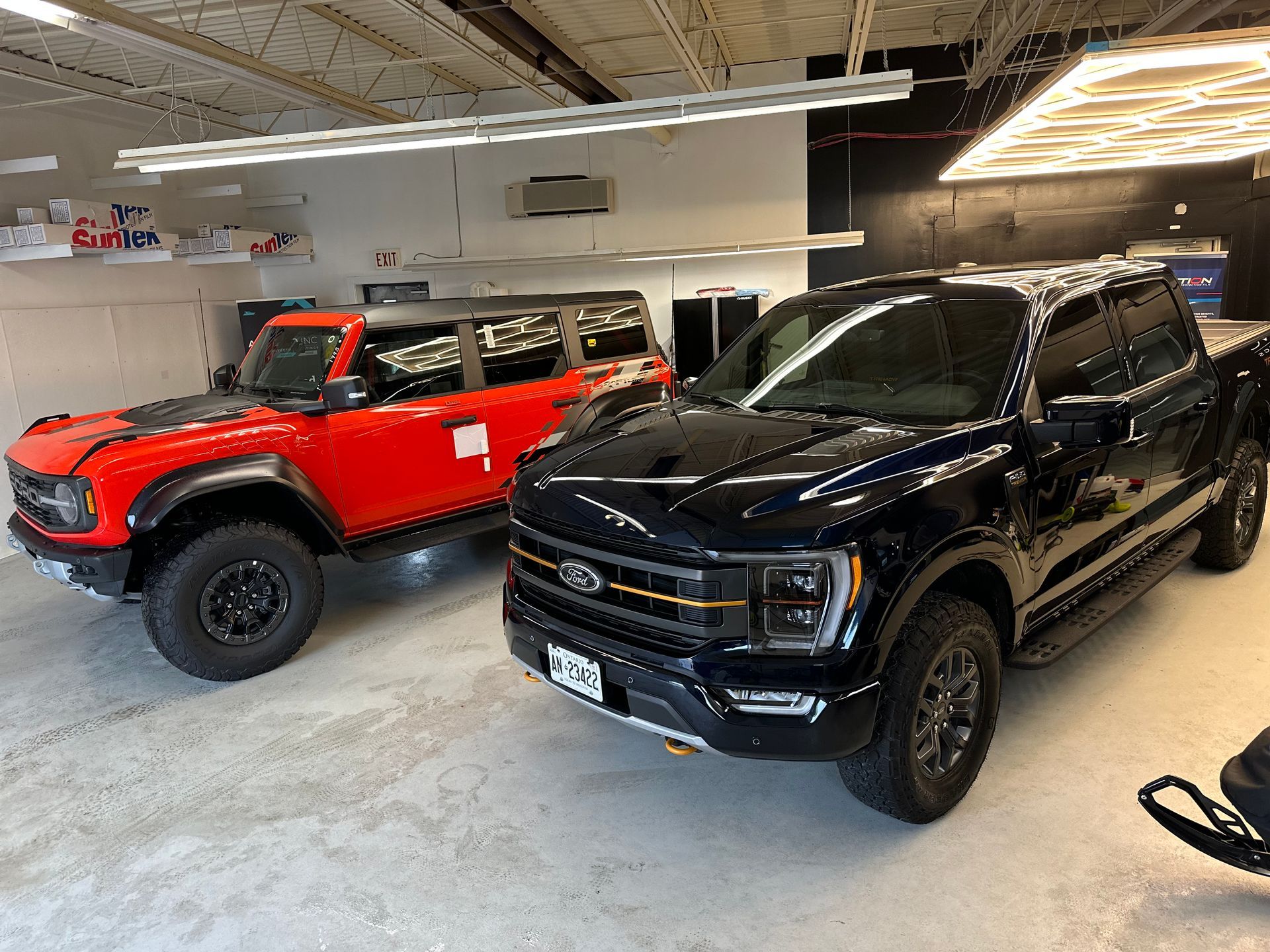 A bright orange Ford Bronco and a black Ford F-150 pickup truck parked inside a bright, clean garage.