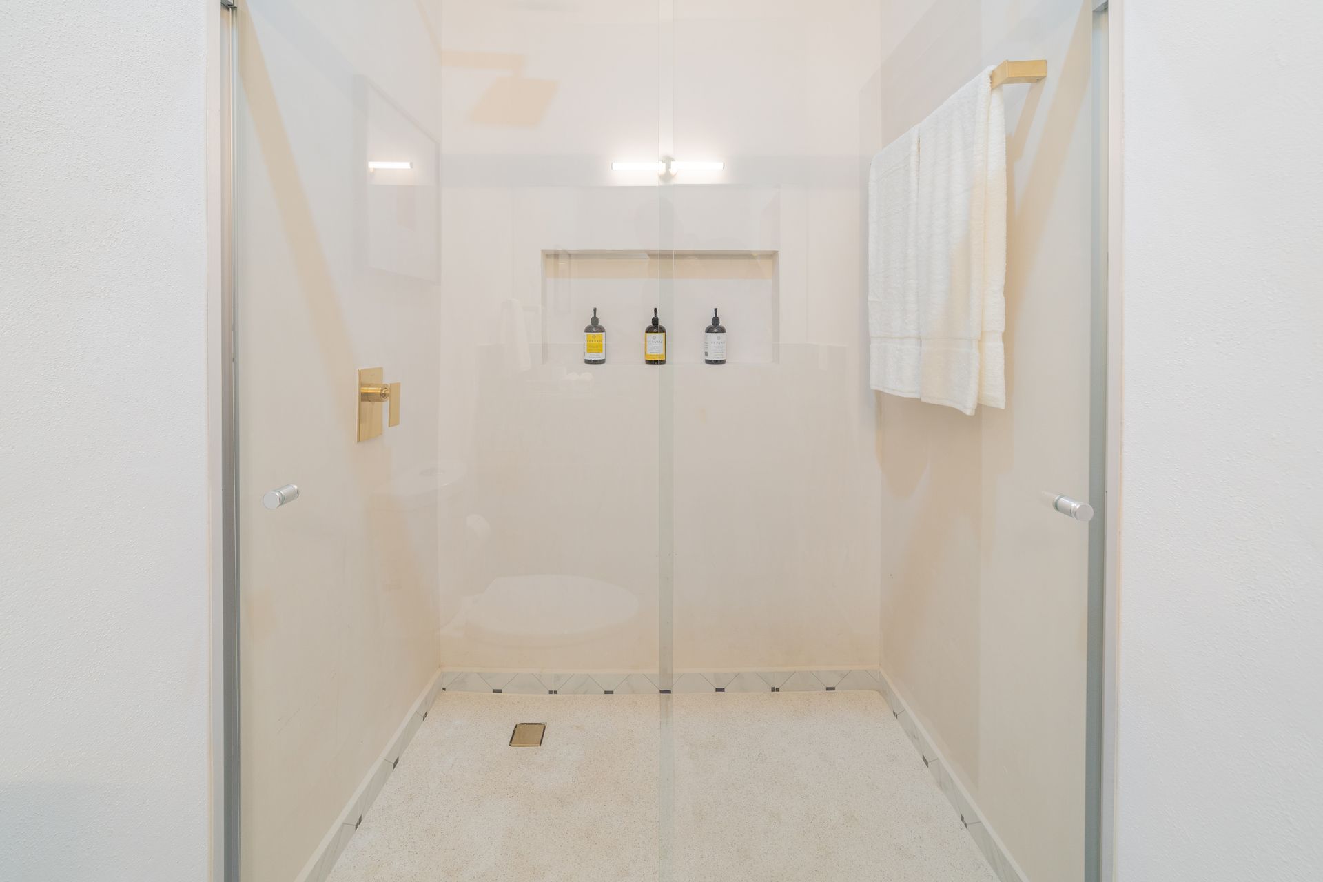 Ducha luminosa, limpia y moderna con puertas de vidrio, piso de baldosas neutrales, estantes empotrados y una toalla en un estante de madera.