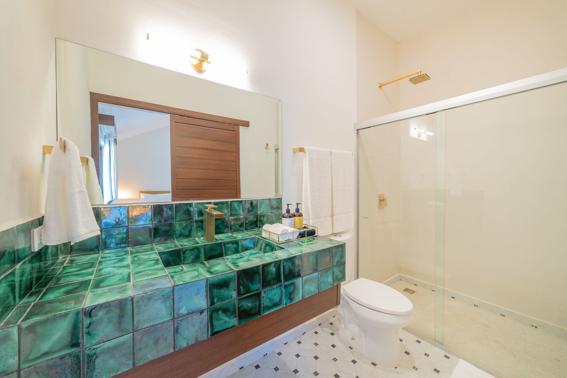 Baño con tocador de azulejos verdes, accesorios dorados, ducha de vidrio y paredes blancas.