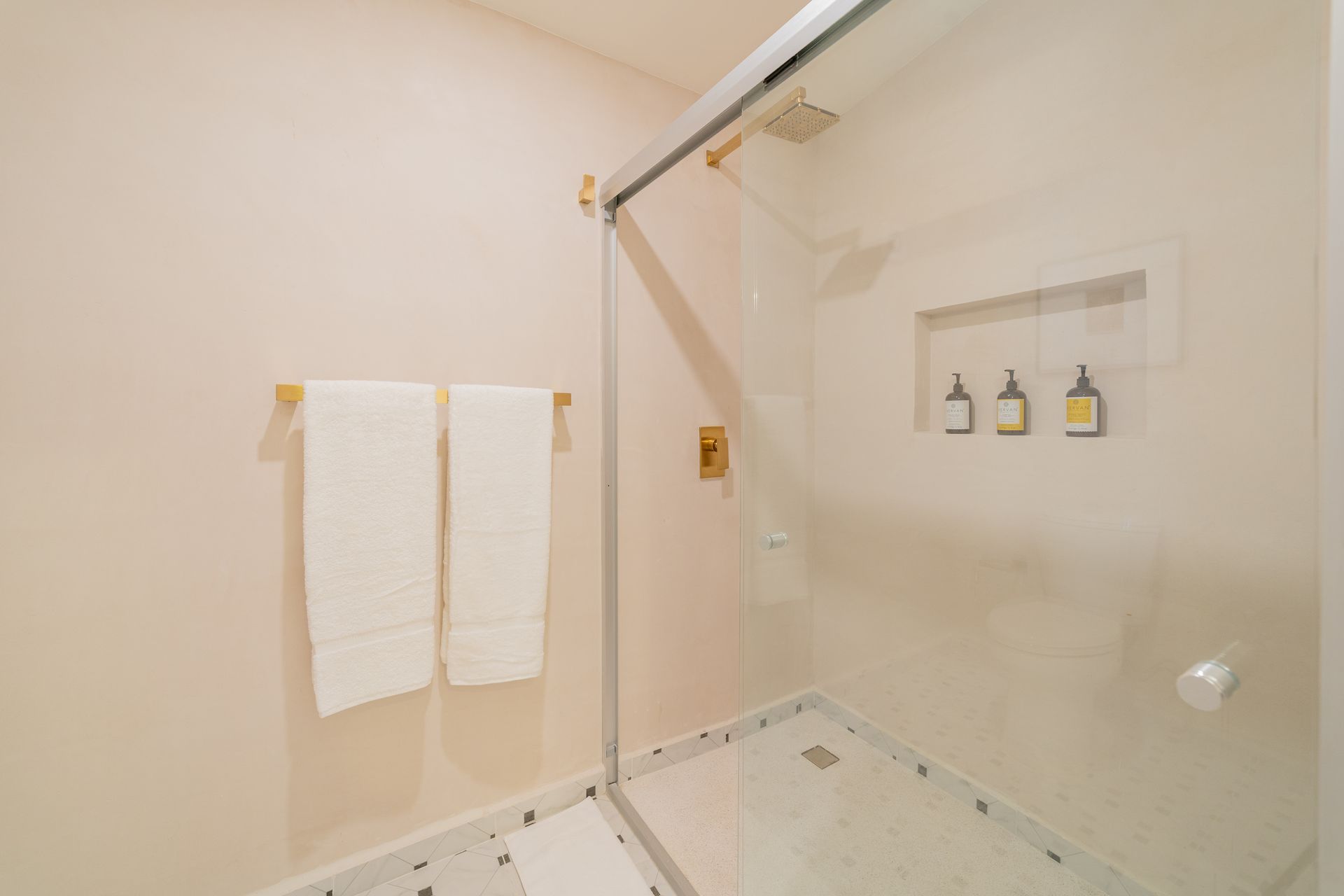 Baño con ducha de cristal, toallas blancas en estante de madera y dispensadores de artículos de tocador incorporados.