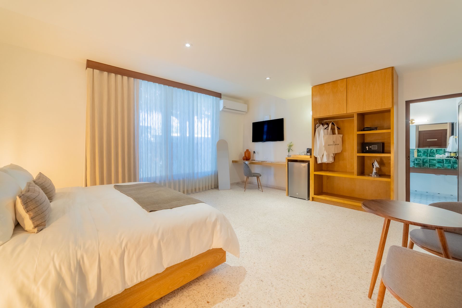 Amplia habitación de hotel con cama, armario, escritorio, televisor y mesa. Los colores claros predominan en el espacio.