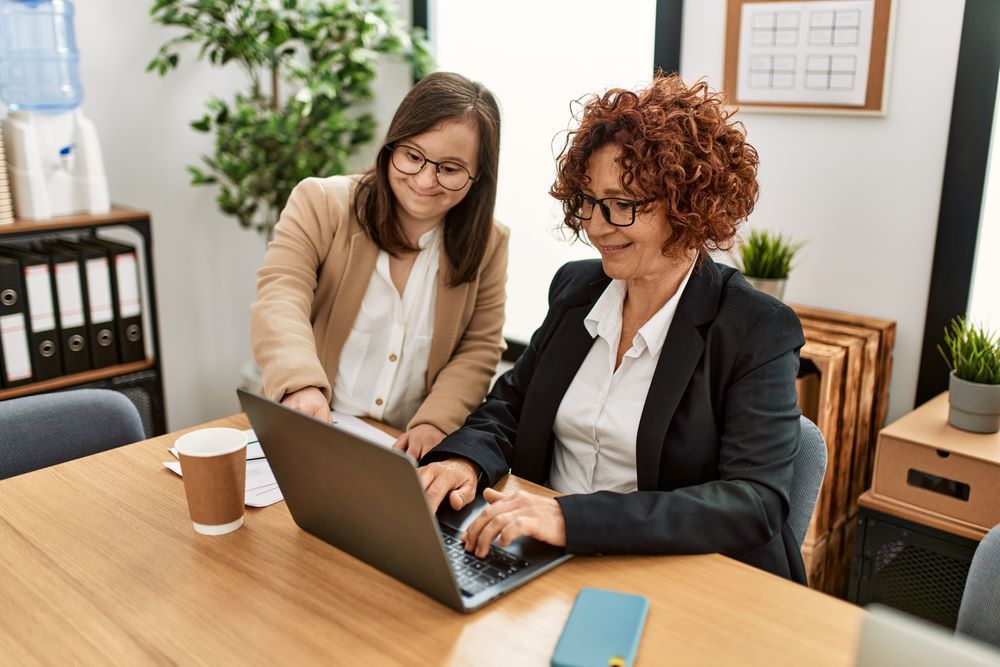 Two Women Are Sitting at a Table Looking at a Laptop Computer — Supportability Care Services in West Rockhampton, QLD
