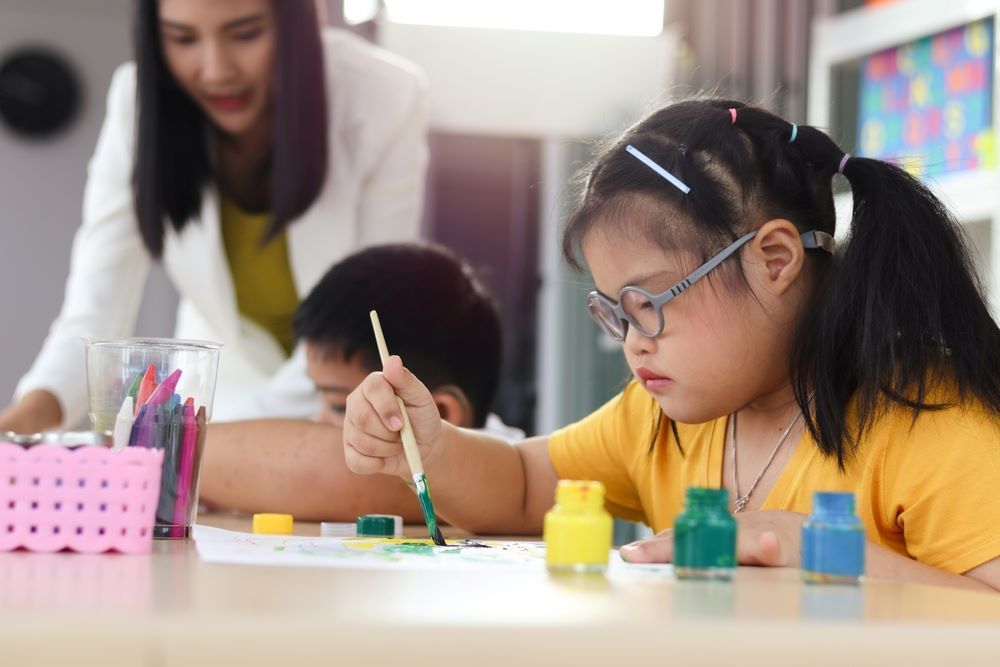 A Girl With Down Syndrome is Painting With a Brush in a Classroom — Supportability Care Services in West Rockhampton, QLD