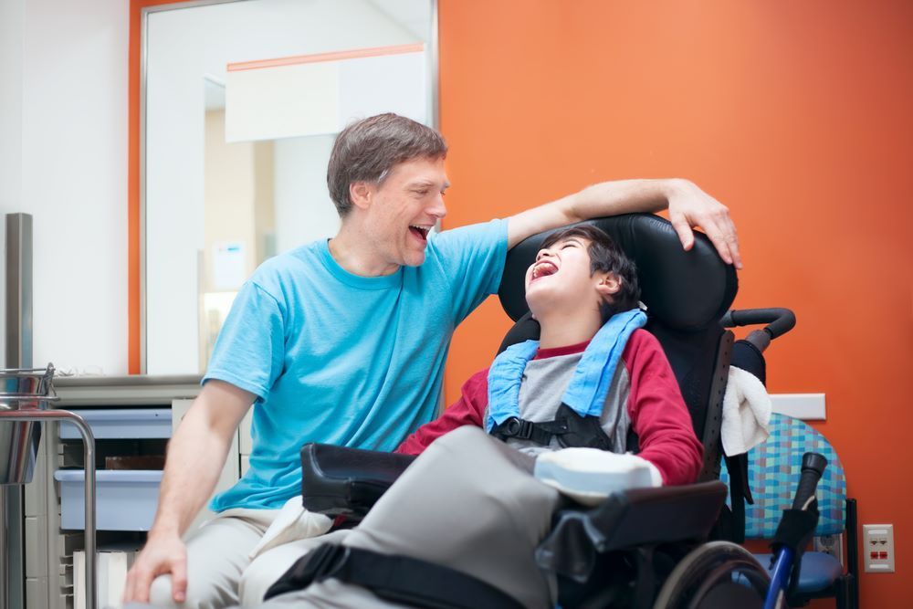 A Man is Sitting Next to a Boy in a Wheelchair — Supportability Care Services in West Rockhampton, QLD