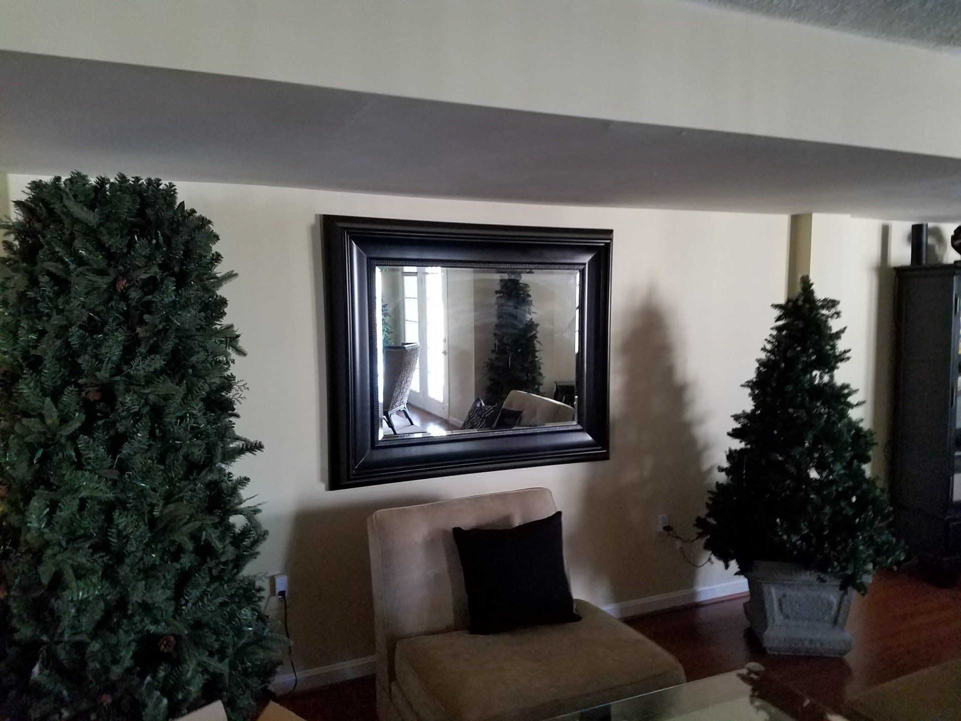 Black-framed mirror centered above a tan armchair with a black pillow. Flanking trees stand beside the chair.