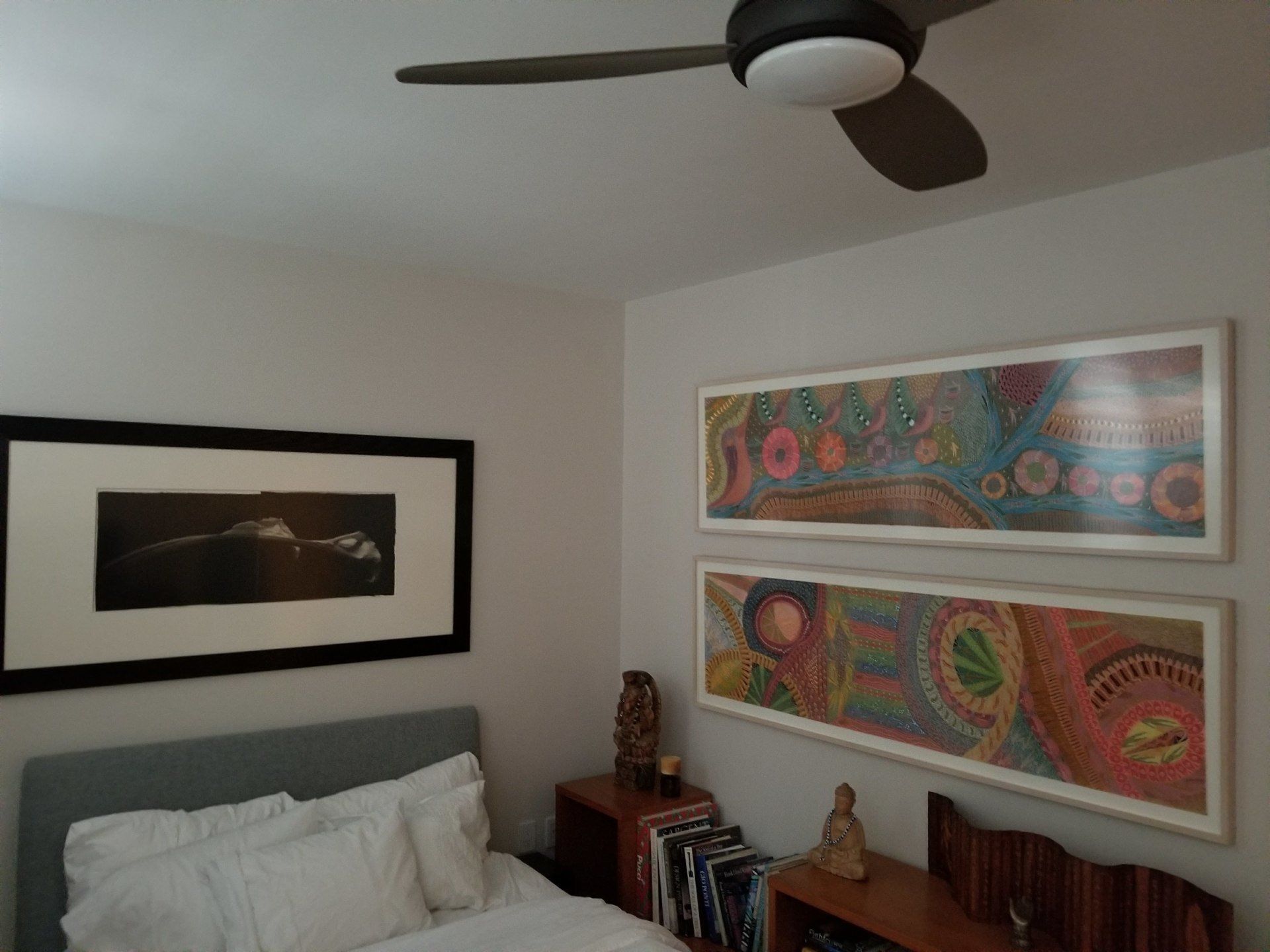 Bedroom with art on the walls, bed, nightstands, ceiling fan, and books.