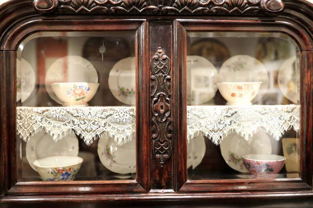 An antique, carved dark wood display cabinet with glass doors containing vintage plates and bowls on lace doilies.