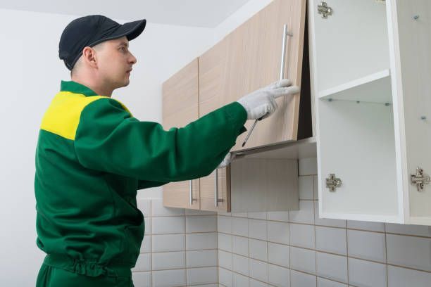 A worker in a green and yellow uniform and white gloves installs a handle on a wooden kitchen cabinet.