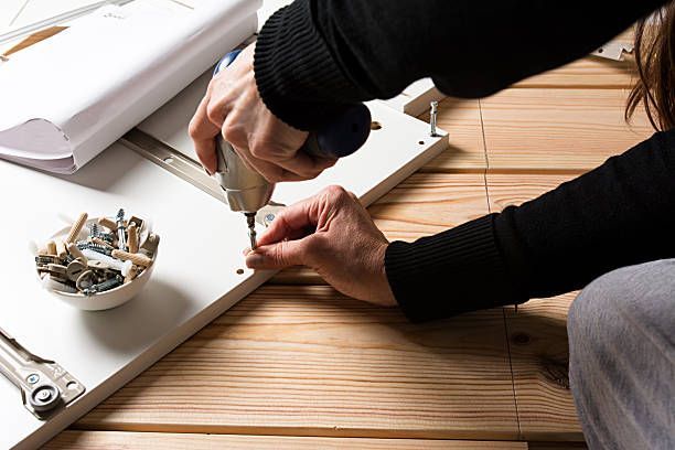Person assembling furniture, using a drill on a white wooden panel, indoors on a wooden floor.