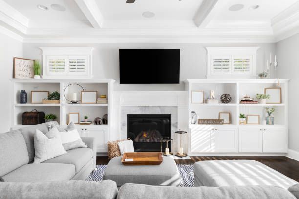 A bright living room with a gray sectional, white built-in shelves flanking a fireplace with a TV mounted above it.