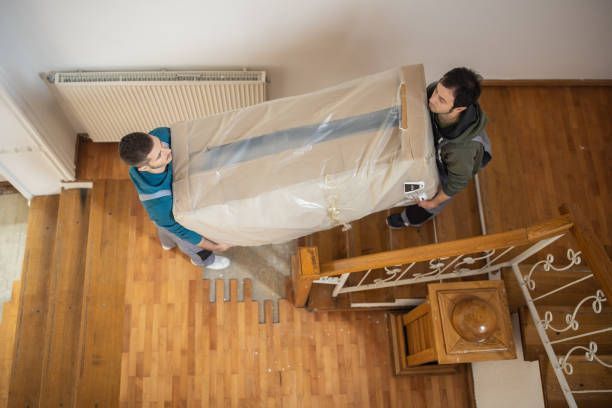 Two people carry a large, plastic-wrapped box down a wooden staircase in an indoor setting.