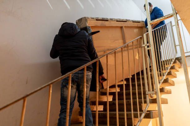 Two people carry a large wooden panel up a staircase in a stairwell.
