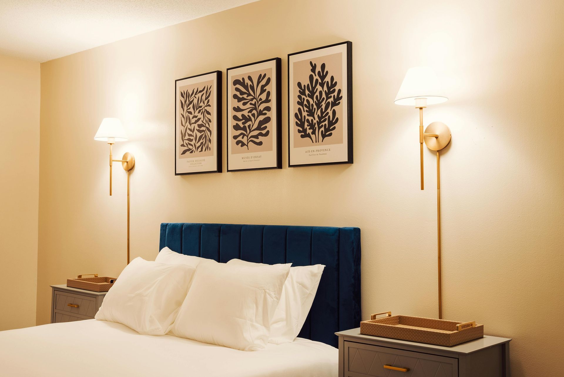 A bedroom with a blue tufted headboard, three framed botanical prints, and two gold wall-mounted lamps above nightstands.