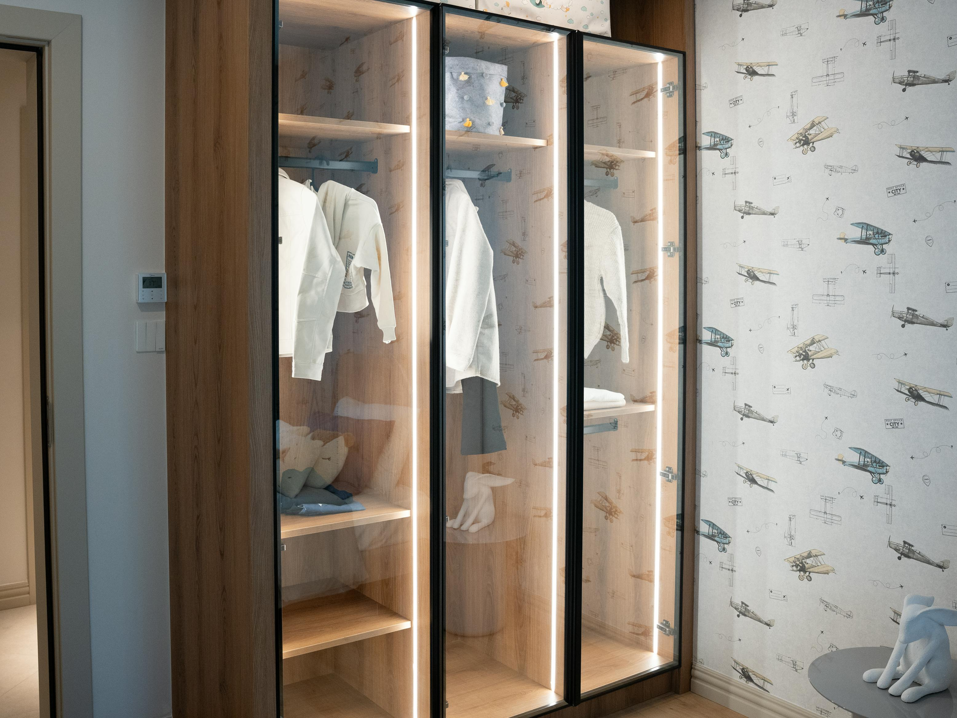 Wooden wardrobe with glass doors and internal lighting, containing clothes and shelves.
