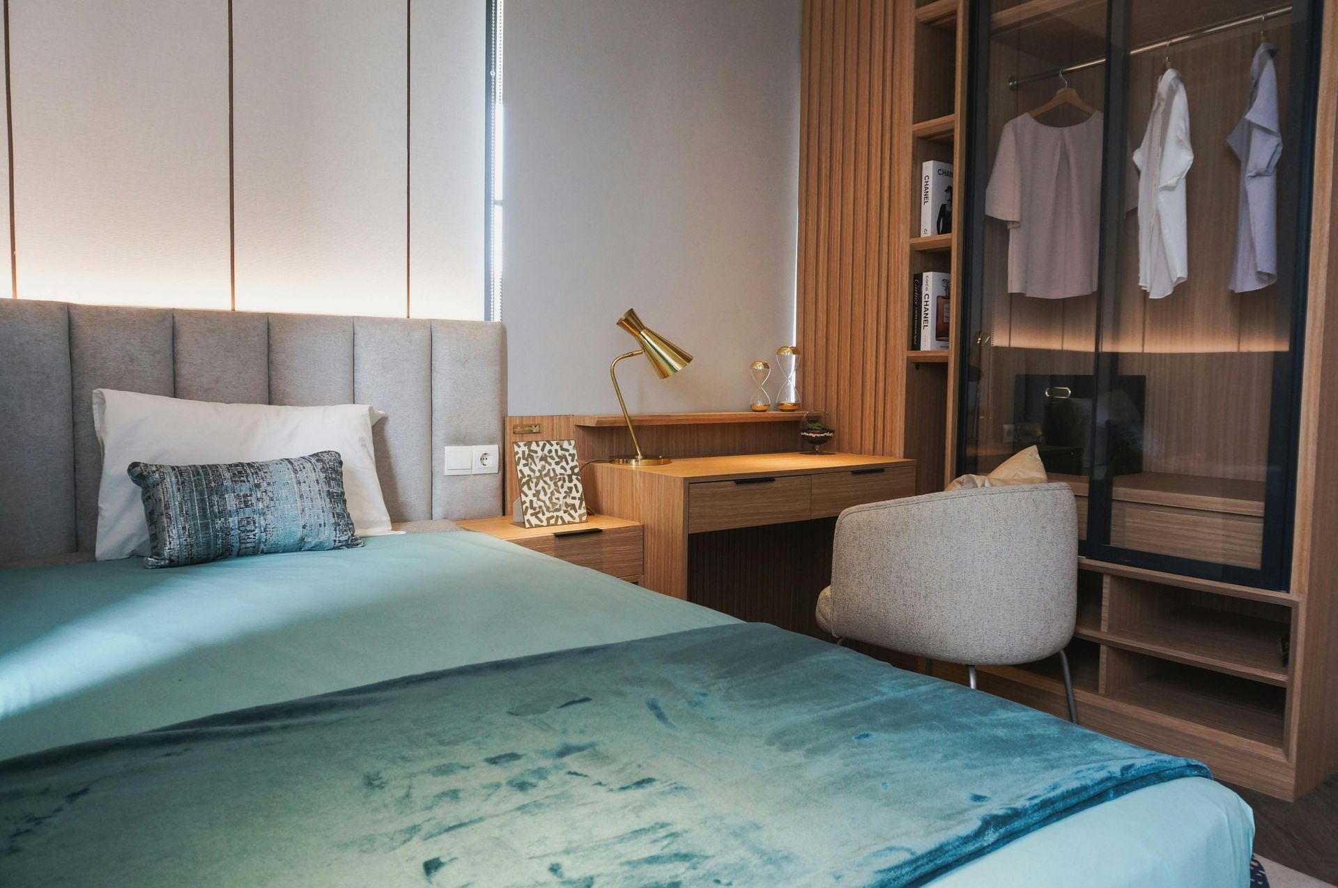 A bedroom with a teal bed, a gray headboard, a wooden desk with a gold lamp, and a glass-front closet with hanging clothes.