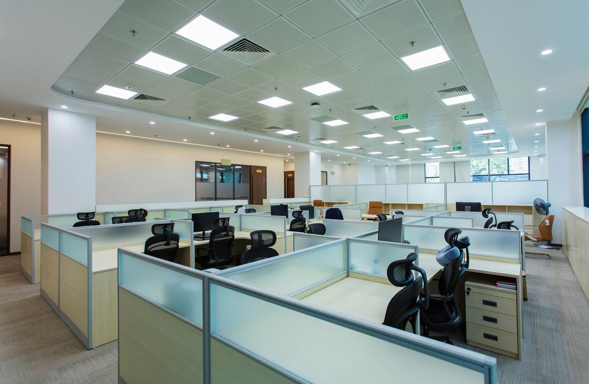Open-plan office with cubicles, desks, computers, and bright fluorescent ceiling lights.