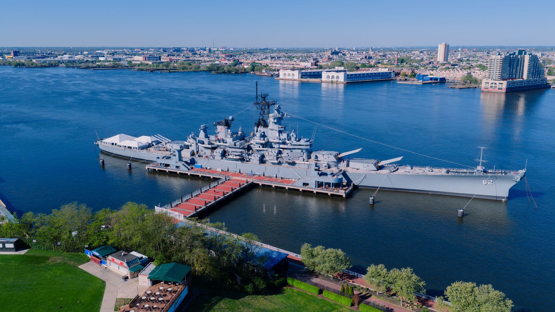 Large gray naval ship docked in a blue harbor with a city skyline in the background