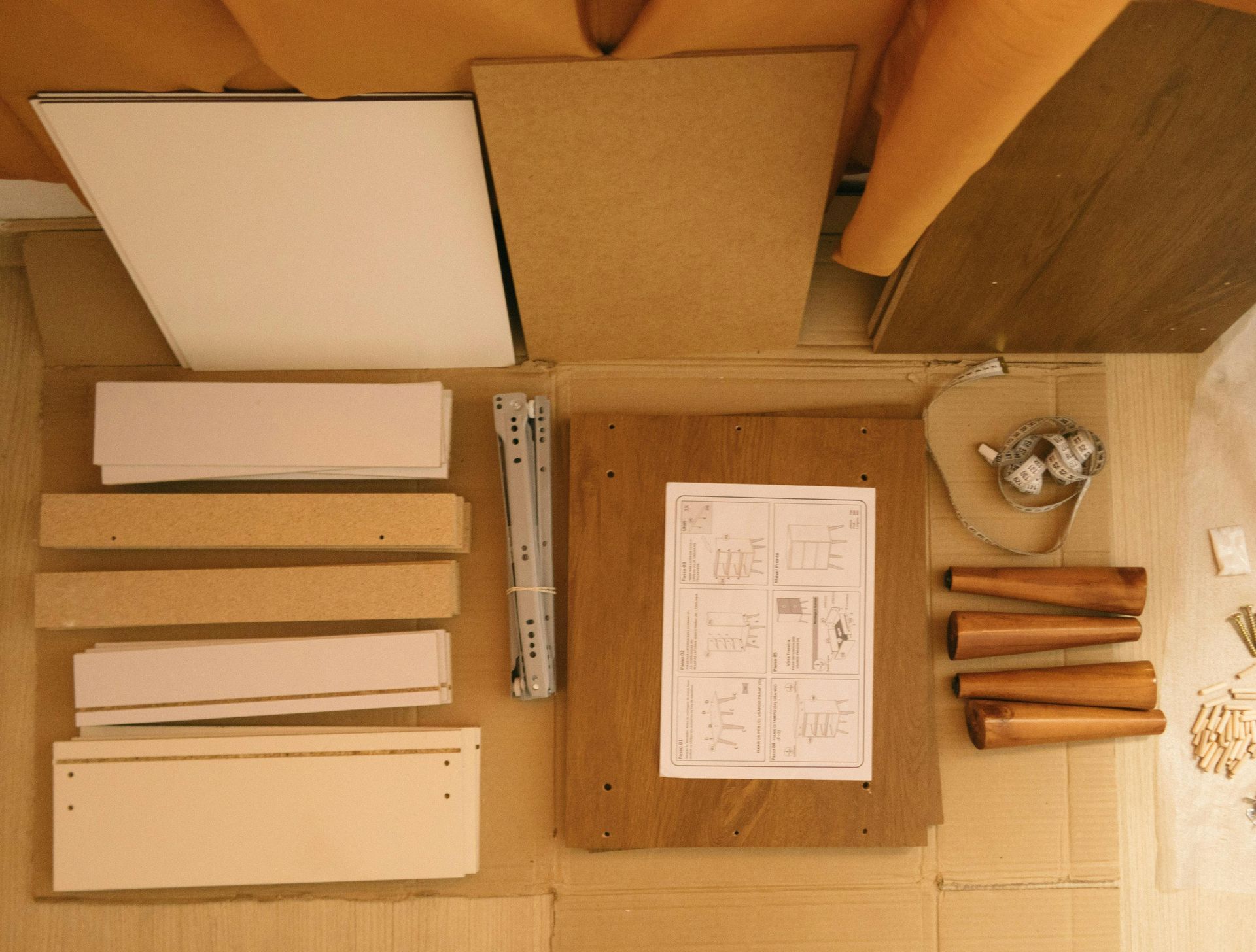 Flat-pack furniture pieces, wooden panels, drawer slides, legs, hardware, and an instruction sheet arranged on cardboard.
