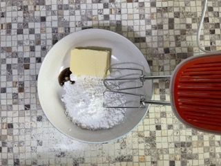 Un piatto con burro, zucchero a velo e un mixer su un bancone piastrellato.