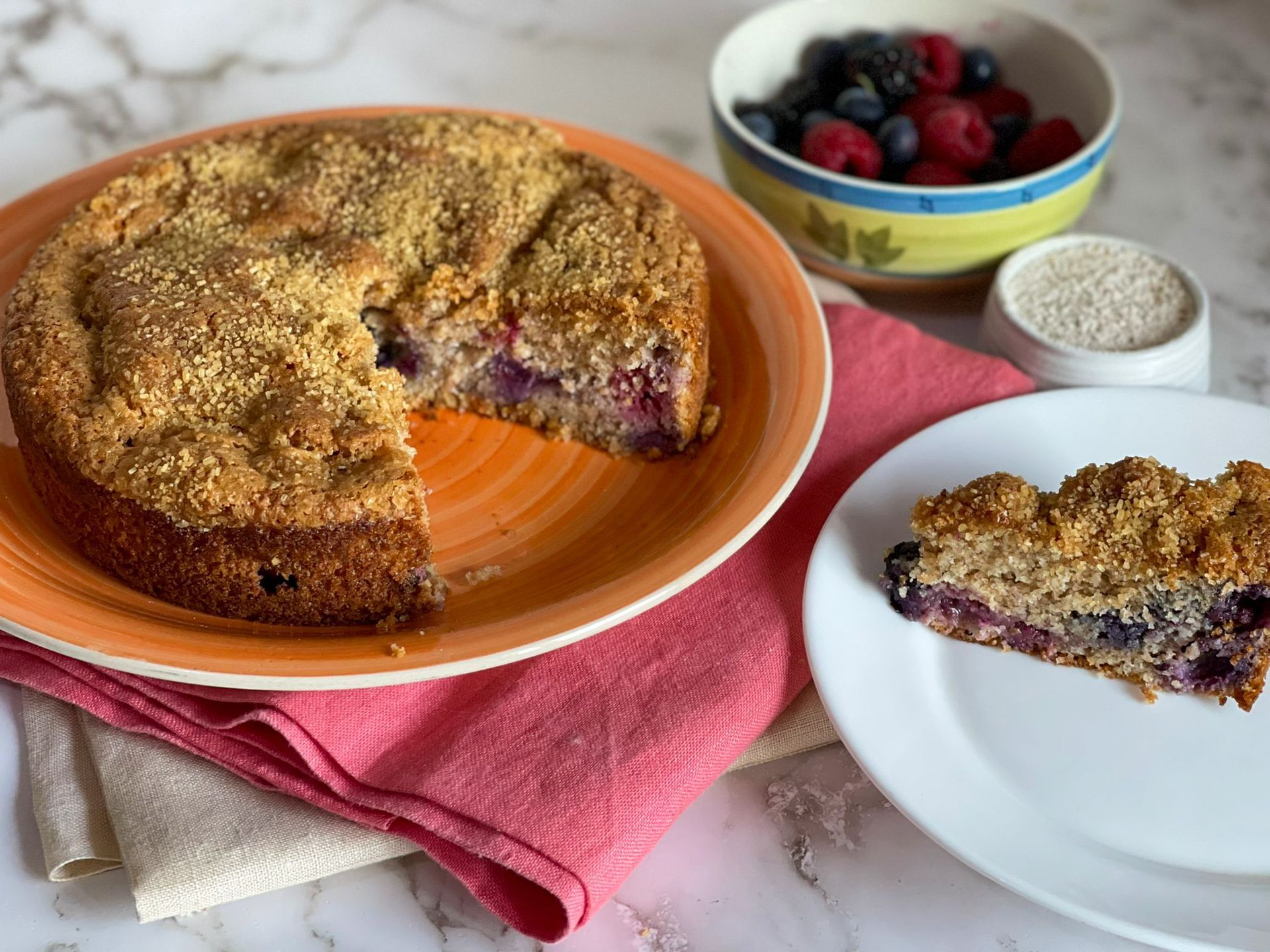 Una torta da cui è stata staccata una fetta si trova su un piatto accanto a una ciotola di frutti di bosco.