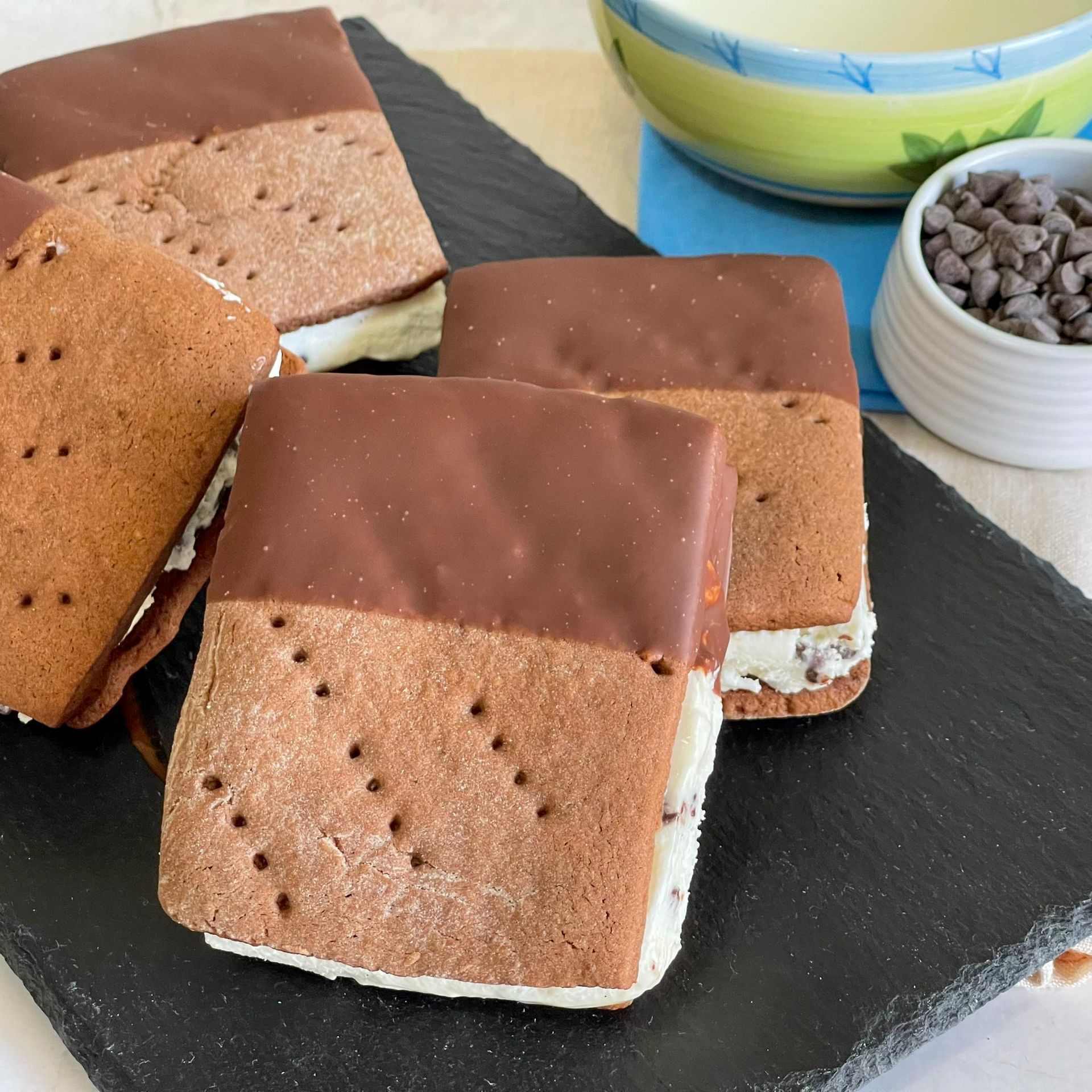 I sandwich al gelato sono appoggiati su un tagliere nero accanto a una ciotola di gocce di cioccolato
