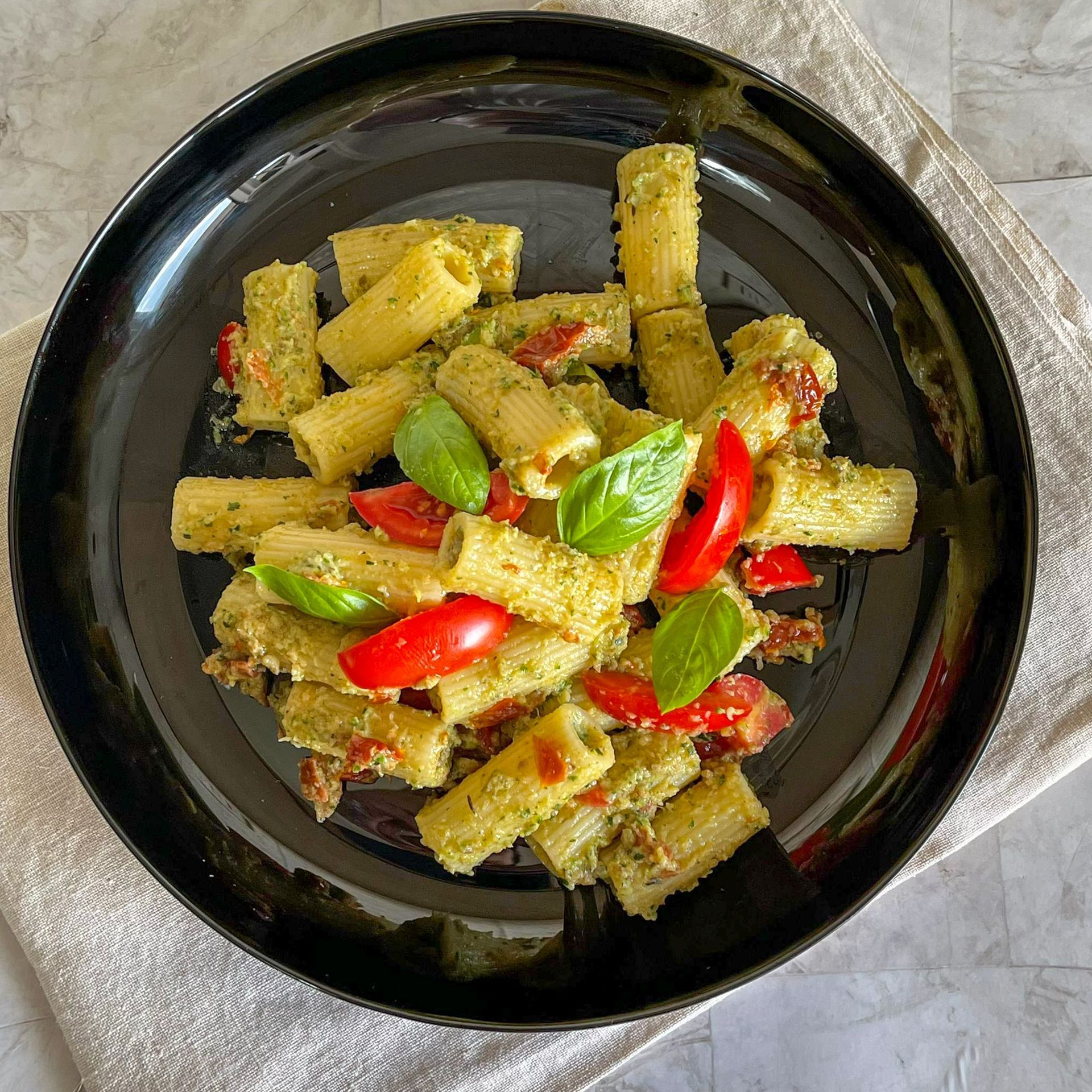 Un piatto nero con sopra pasta, pomodori e basilico su un tavolo.