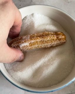 Una persona tiene in mano un churro in una ciotola di zucchero.