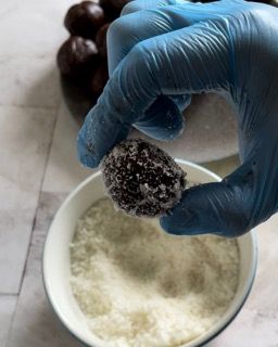Una persona che indossa guanti blu tiene in mano un tartufo al cioccolato sopra una ciotola di zucchero a velo.