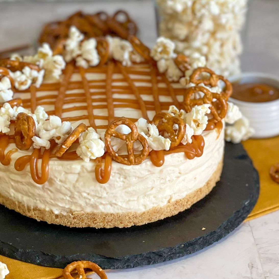 Una cheesecake con popcorn e pretzel sopra è su un piatto nero.