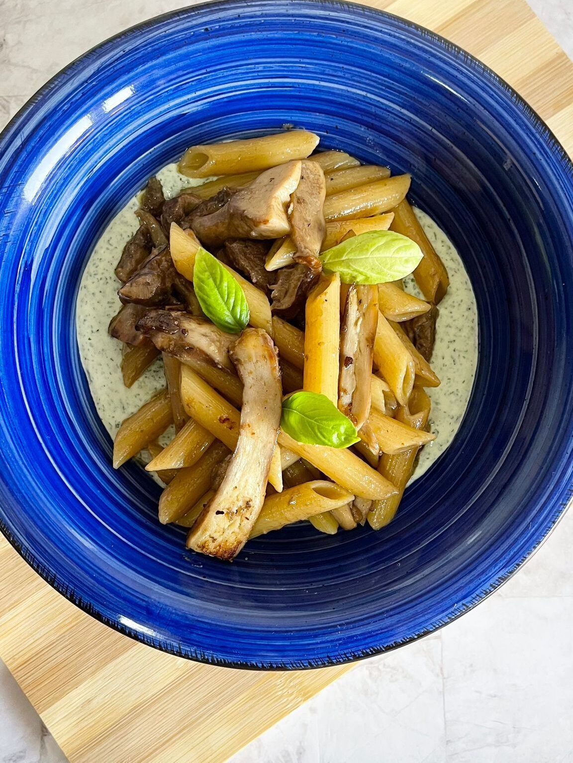 Un piatto blu condito con pasta, funghi e basilico su un tagliere di legno.