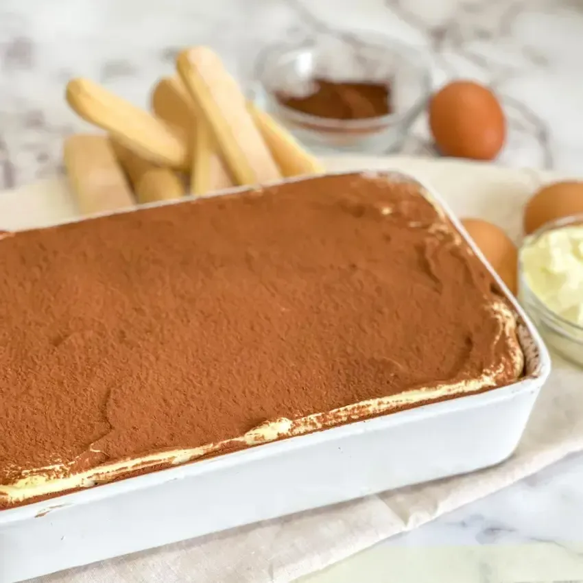 Tiramisù classico fatto in casa in una teglia di ceramica e un piatto, con una generosa spolverata di cacao amaro. 