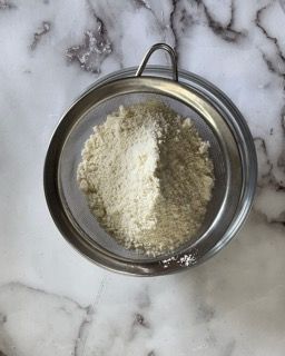 Una ciotola di farina viene setacciata attraverso un colino su un tavolo.