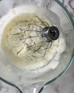 Una ciotola di vetro piena di panna montata e una frusta.