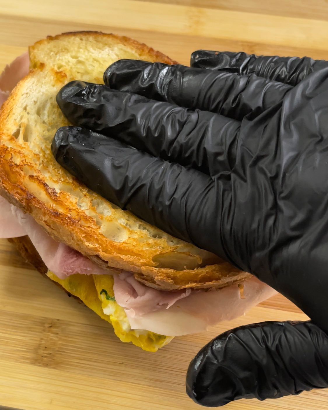 Una persona che indossa guanti neri tiene in mano un panino al prosciutto e all'uovo.