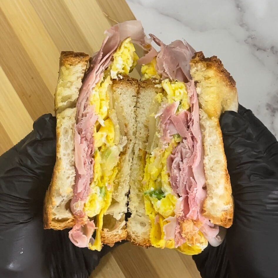 Una persona che indossa guanti neri tiene in mano un panino al prosciutto e all'uovo.