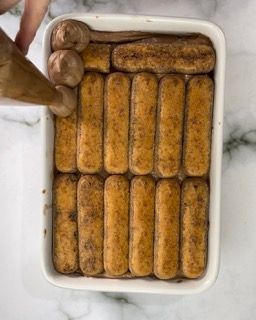 Una persona versa la salsa al cioccolato su una teglia piena di bastoncini di tiramisù.