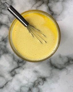 Una ciotola di liquido giallo con una frusta al suo interno su una superficie di marmo.