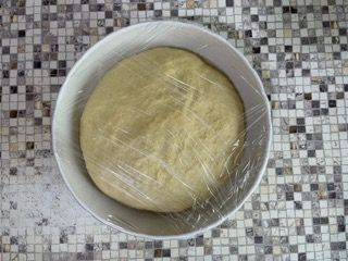 Una ciotola di impasto è avvolta nella pellicola trasparente su un tavolo piastrellato.