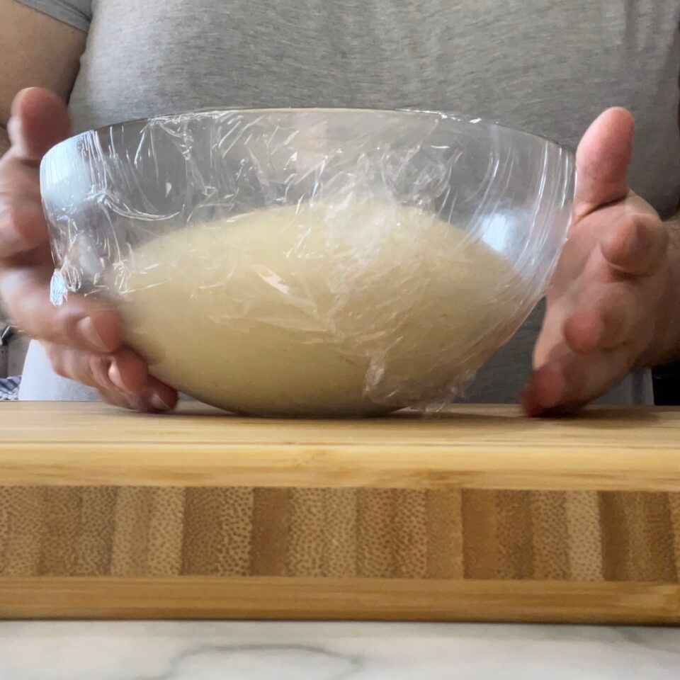 Una persona tiene in mano una ciotola di impasto avvolta nella pellicola trasparente.